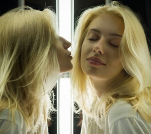 Das Bild verdeutlicht ein Prinzip der Reinkarnationstherapie: Umwelt als Spiegel. Eine blonde Frau gibt ihrem Spiegelbild einen Kussmund, während dieses eine Wange hinhält.