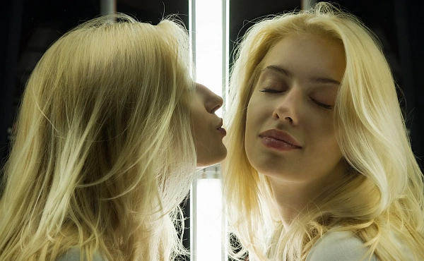 Eine junge blonde Frau küsst ihr Spiegelbild, während dieses ihr eine Wange hinhält. Es steht symbolisch für das Prinzip Umwelt als Spiegel.