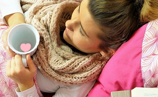 Frau liegt warm eingehüllt und hält eine Tasse mit Herzsymbol – Sinnbild für liebevolle Selbstfürsorge und die seelische Begleitung bei Krebs in der Reinkarnationstherapie.